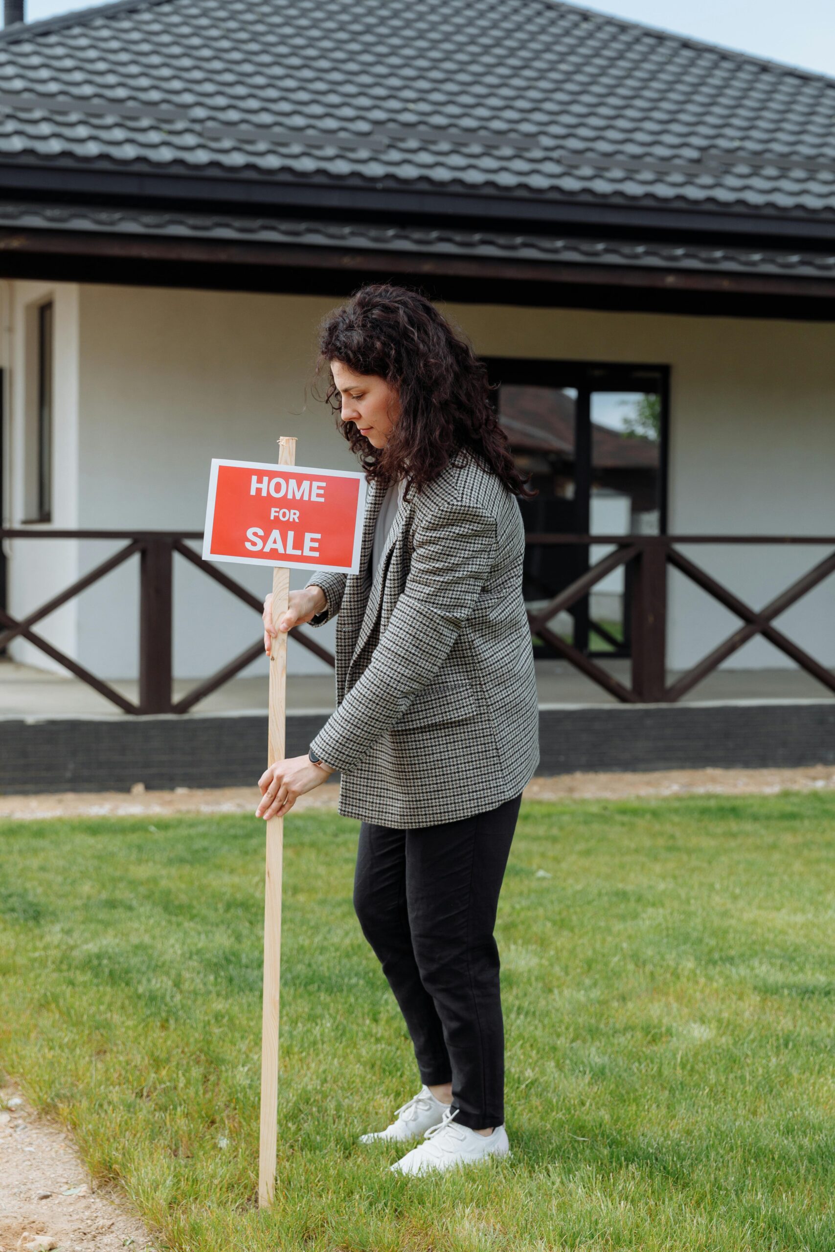 Woman placing a home for sale sign in front of a house, showcasing real estate career.
